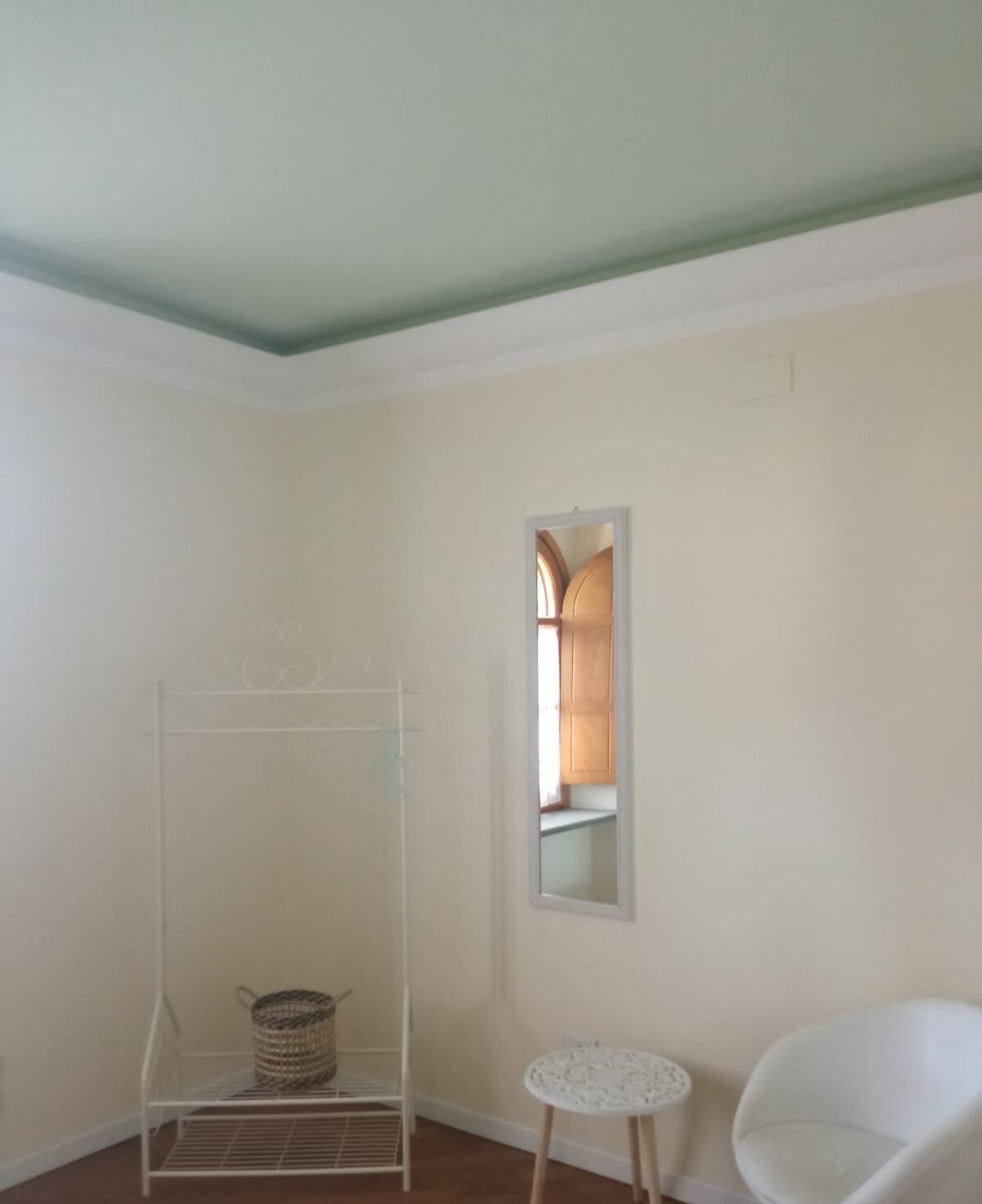 Una habitación con una silla blanca, una pequeña mesa redonda, una estantería metálica y un espejo en la pared.