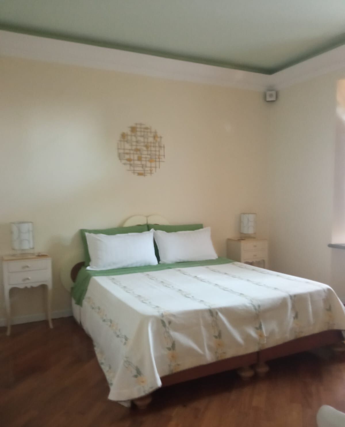 A bedroom with a bed, green headboard, white bedding, wooden floor, bedside tables, and a wall decoration.