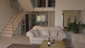 Un salón espacioso con un sofá color beige, una mesa de centro con copas de vino y fruta, una escalera y vistas al área de la cocina y el comedor.
