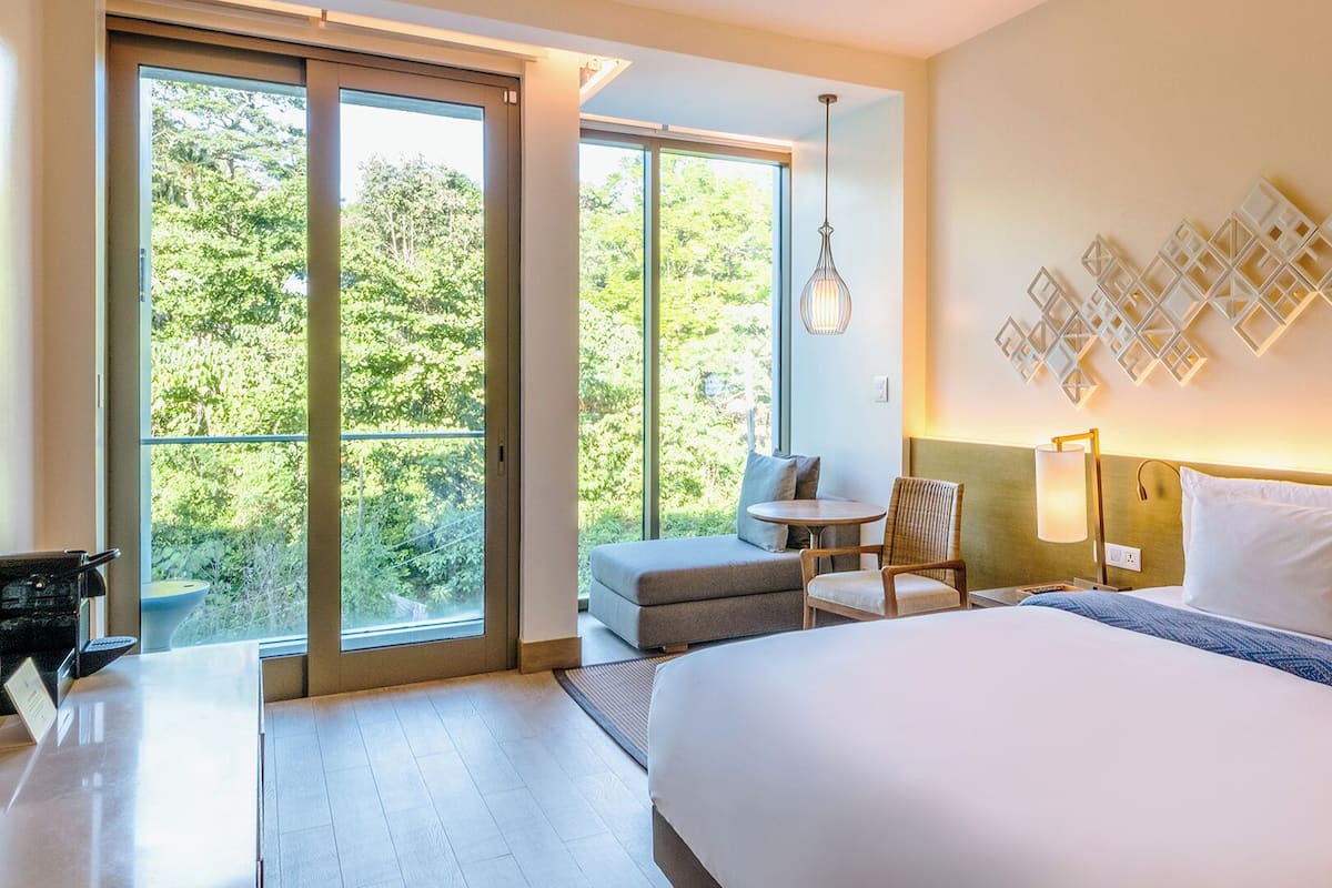 Une chambre d’hôtel dotée d’un grand lit, d’un coin salon avec un fauteuil et une petite table, d’un bureau avec un ordinateur, et offrant une vue sur la verdure par les fenêtres.