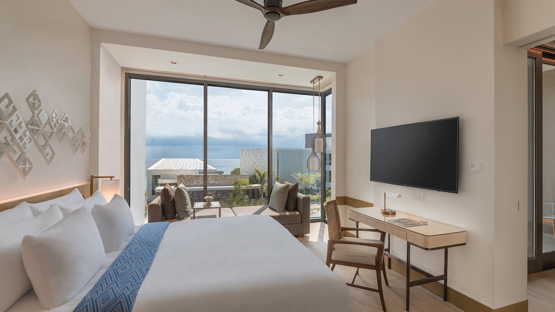 A spacious hotel room with a large bed, a desk, a flat-screen TV, and a balcony with a view of the ocean.