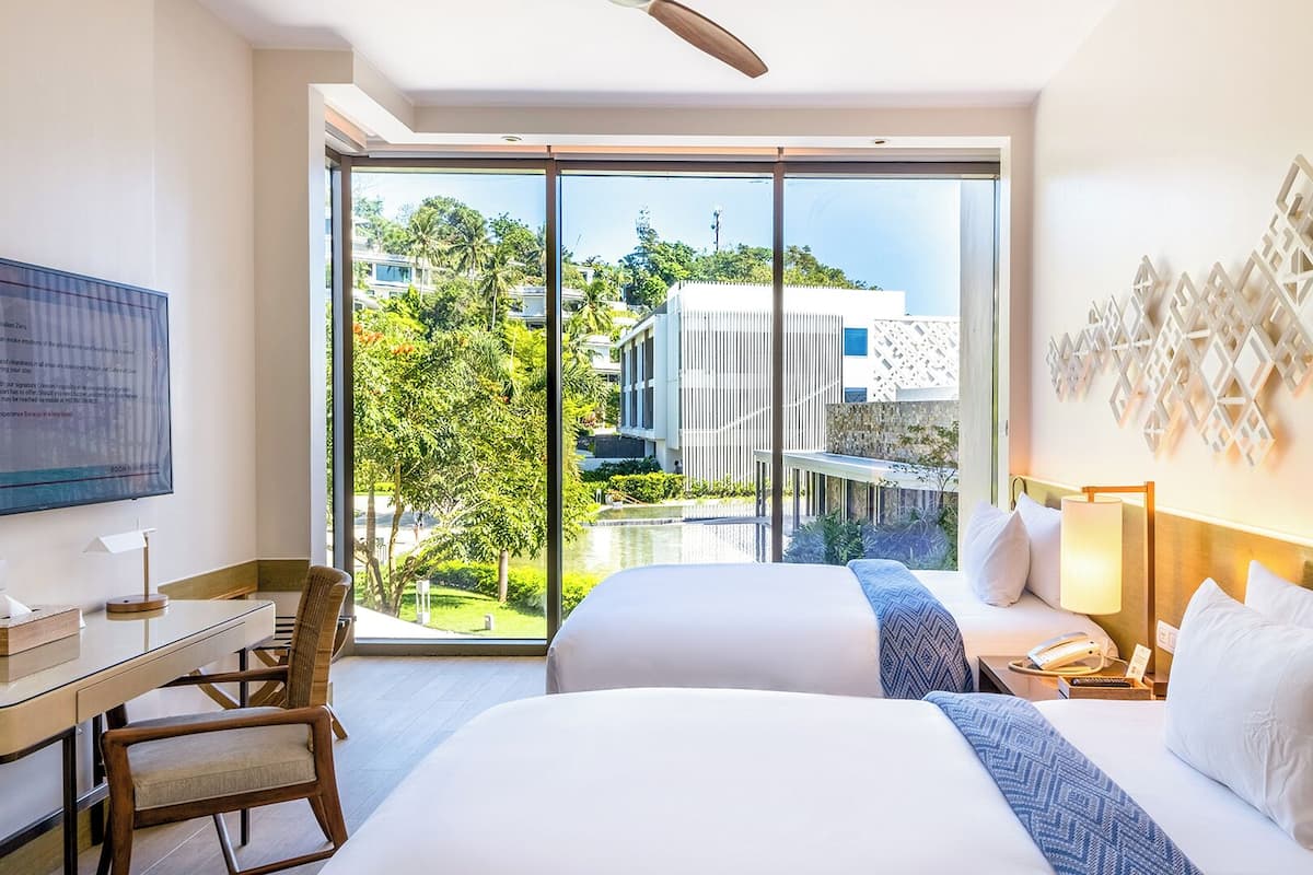Une chambre d’hôtel avec un grand lit, un bureau et une chaise. Il y a un téléviseur accroché au mur et une fenêtre donnant sur des immeubles et des arbres.