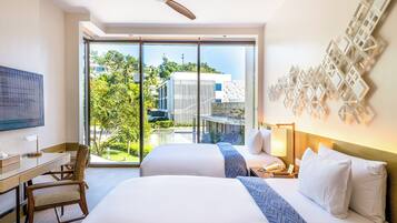 A hotel room with a large bed, a desk, and a chair. There is a TV on the wall and a window with a view of buildings and trees.