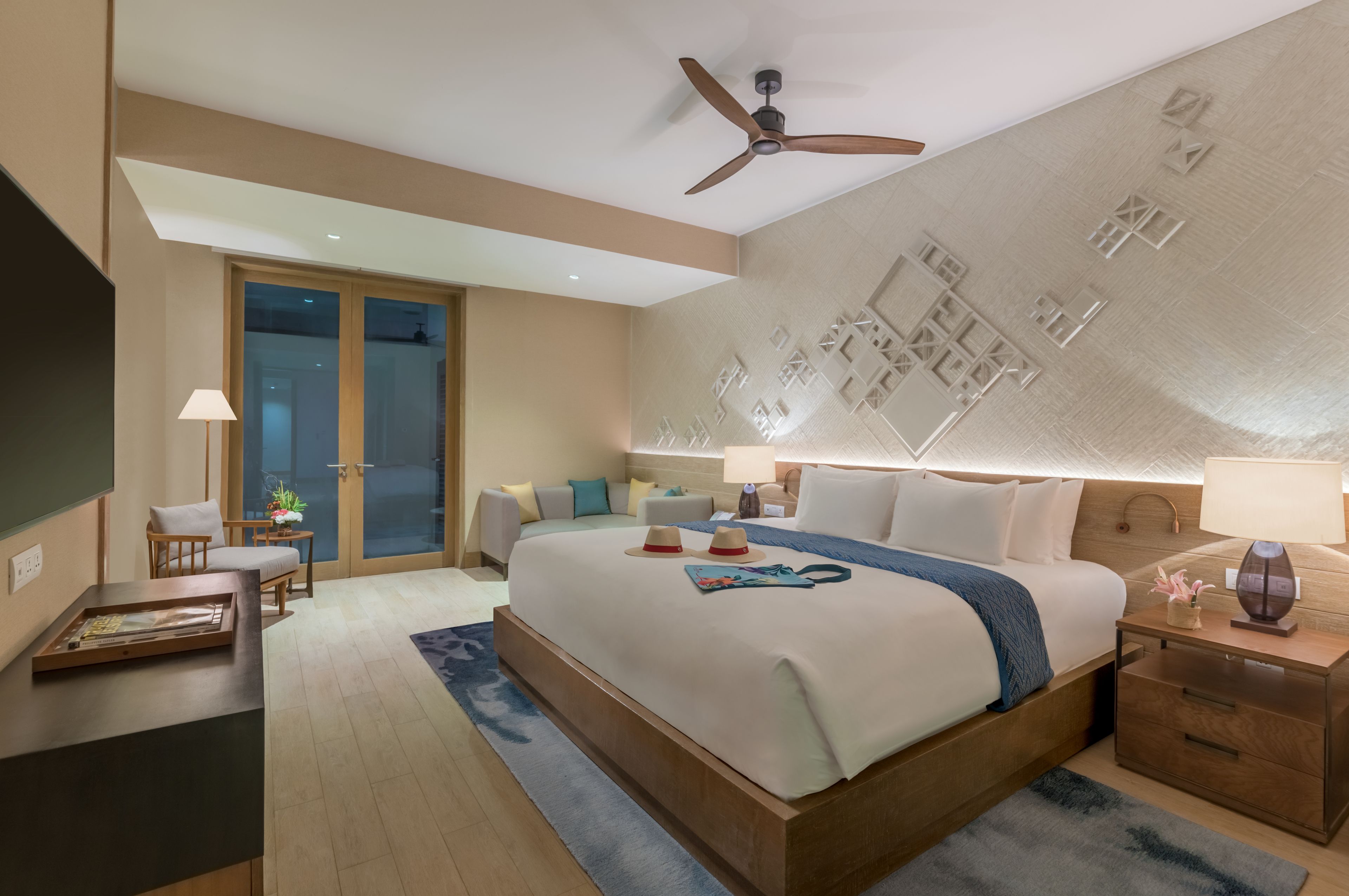 A modern hotel room with a large bed, a ceiling fan, a nightstand, a desk, and a view of the outside through a glass door.