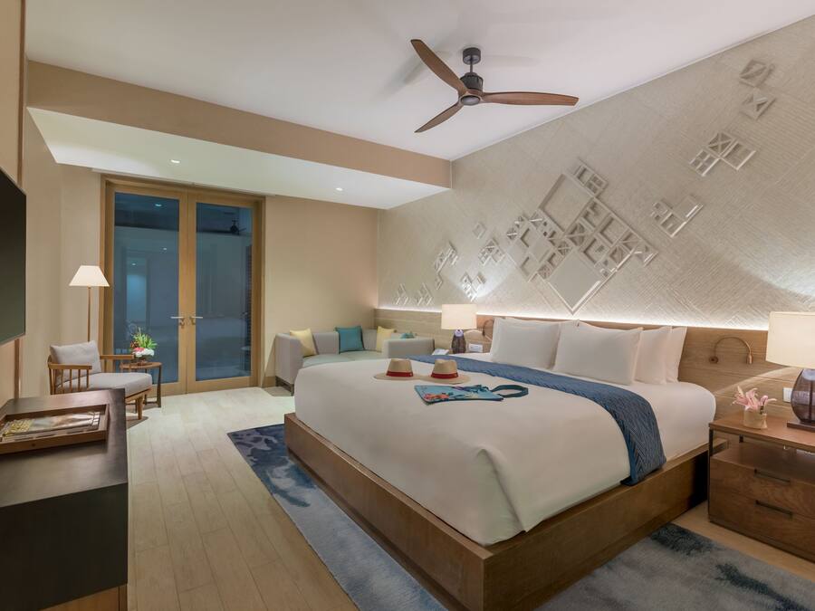 A modern hotel room with a large bed, a ceiling fan, a nightstand, a desk, and a view of the outside through a glass door.