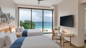 Une chambre d’hôtel avec deux lits, un bureau, une chaise, une télévision et une grande fenêtre donnant sur l’océan et les arbres.