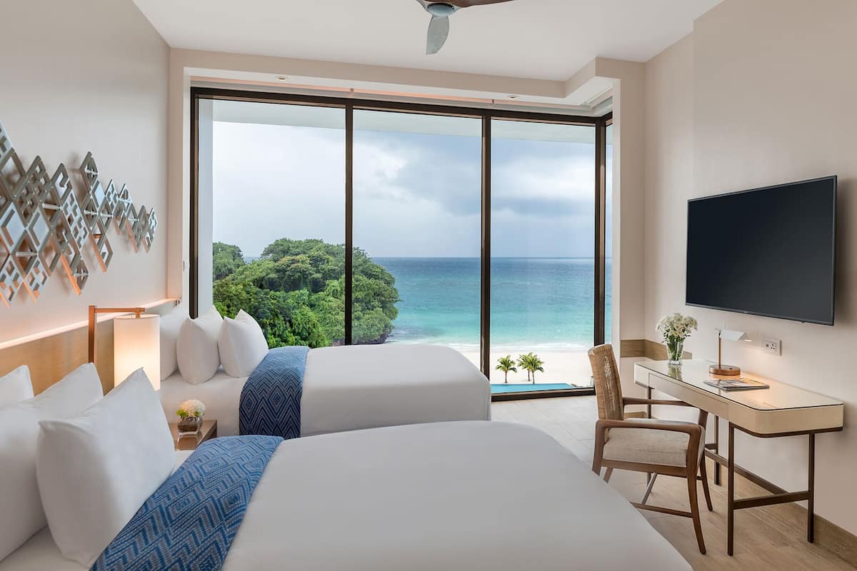 Une chambre d’hôtel avec deux lits, un bureau, une chaise, une télévision et une grande fenêtre donnant sur l’océan et les arbres.