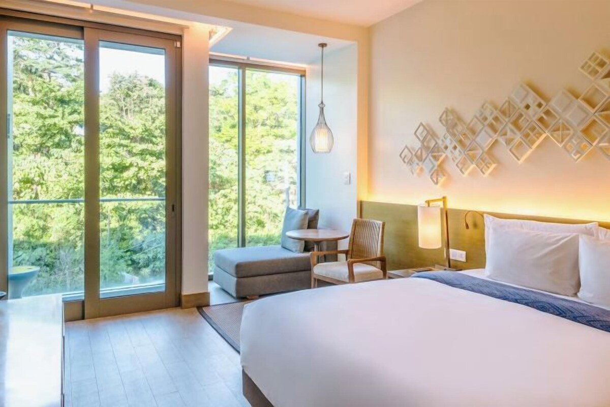 Une chambre d’hôtel dotée d’un grand lit, d’un coin salon avec une table et offrant une vue sur la verdure par les fenêtres.