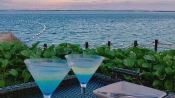 Dos copas de martini sobre una mesa trenzada, con vista al mar y una costa lejana.