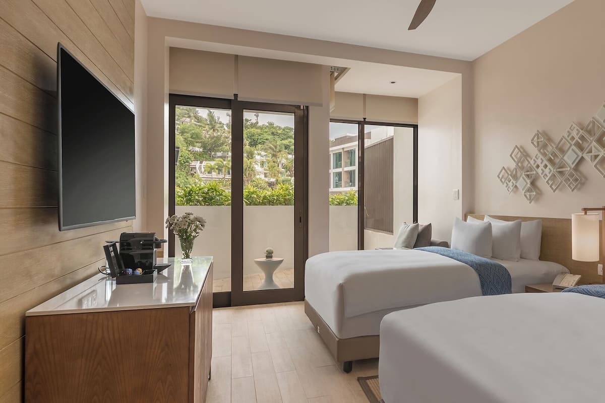 Une chambre d’hôtel avec deux lits, un bureau, un téléviseur à écran plat et un balcon offrant une vue sur la verdure et les bâtiments.