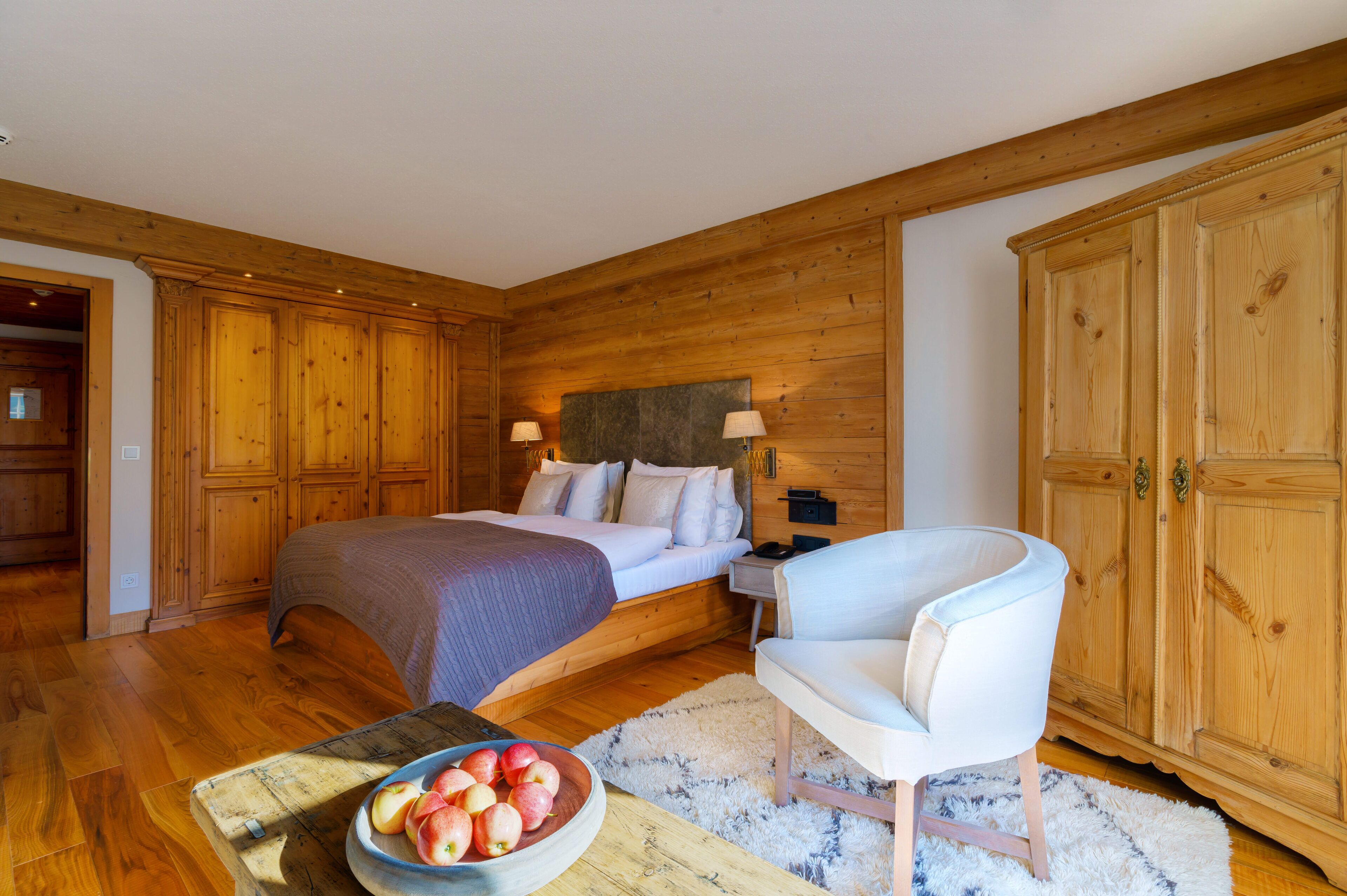 Ein Schlafzimmer mit Holzvertäfelung, einem Bett mit weißen Leinen, einem Nachttisch, einem Sessel und einer Obstschale auf einem Tisch.