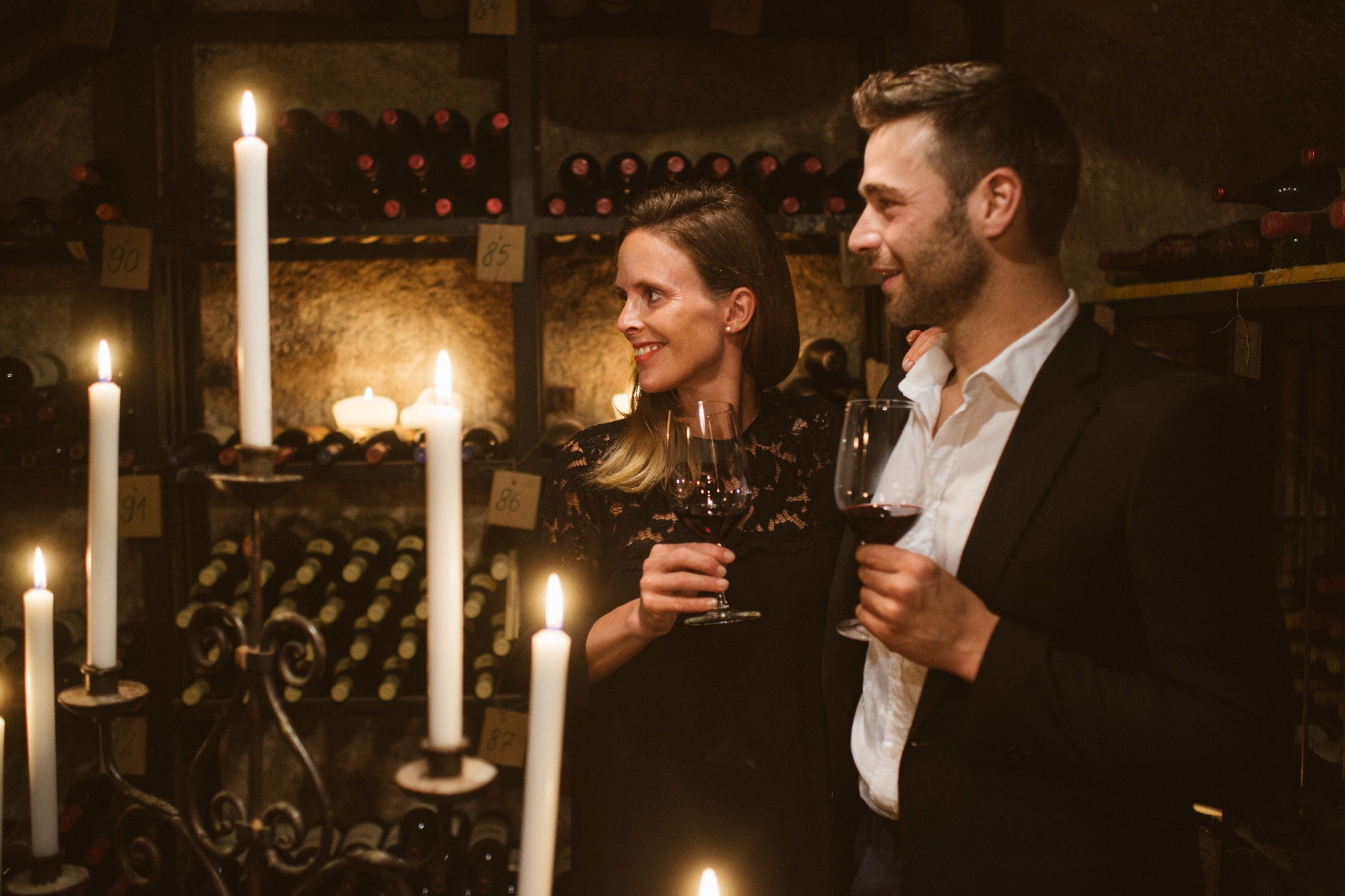 A couple holding wine glasses in a dimly lit room with candles and wine bottles in the background.