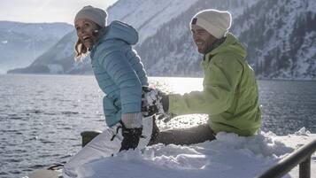 Two people in winter clothing sitting on a snow-covered surface with a body of water and mountains in the background.
