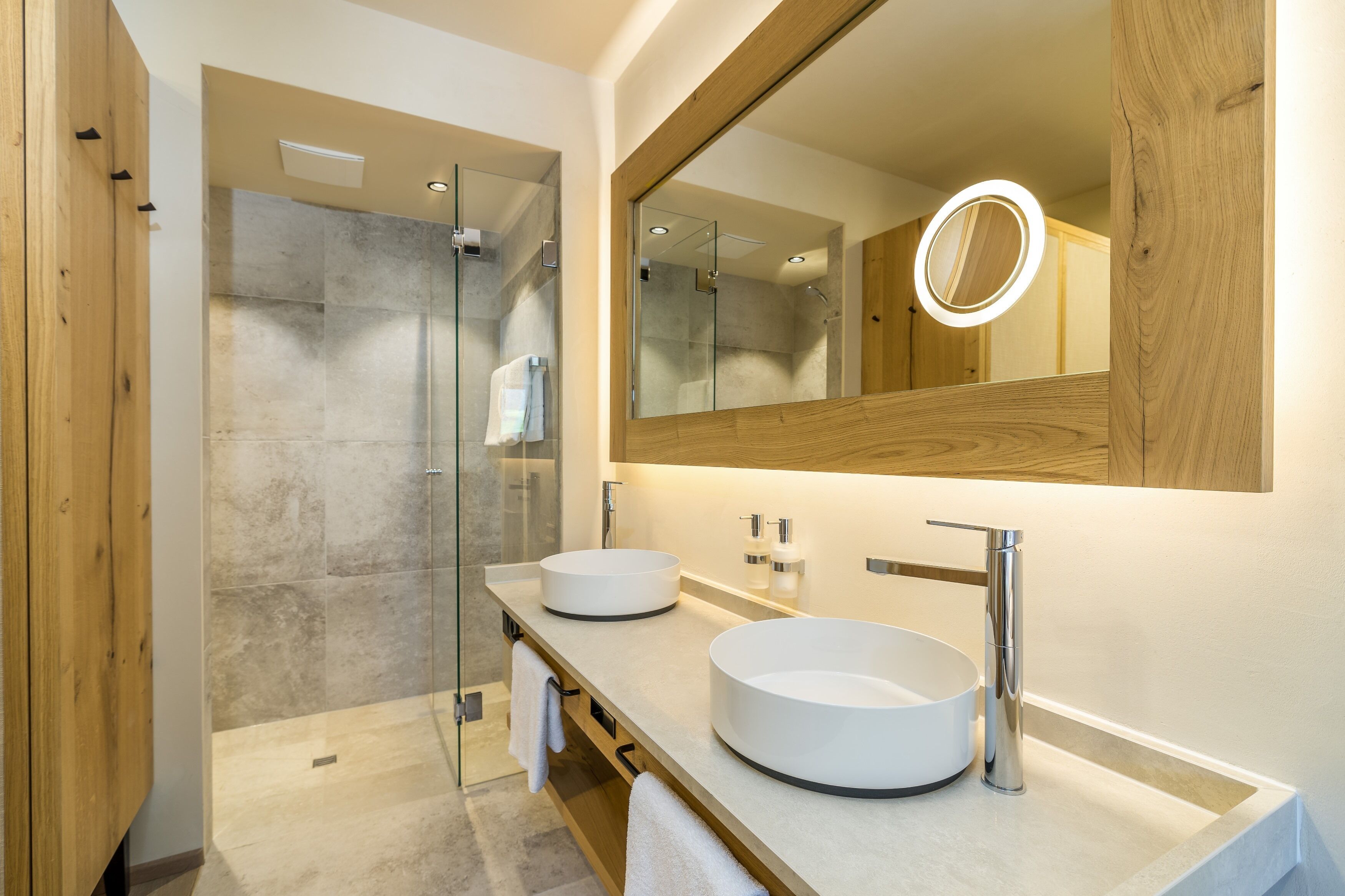 A modern bathroom with two round sinks, a glass shower enclosure, and a large mirror with a circular light.