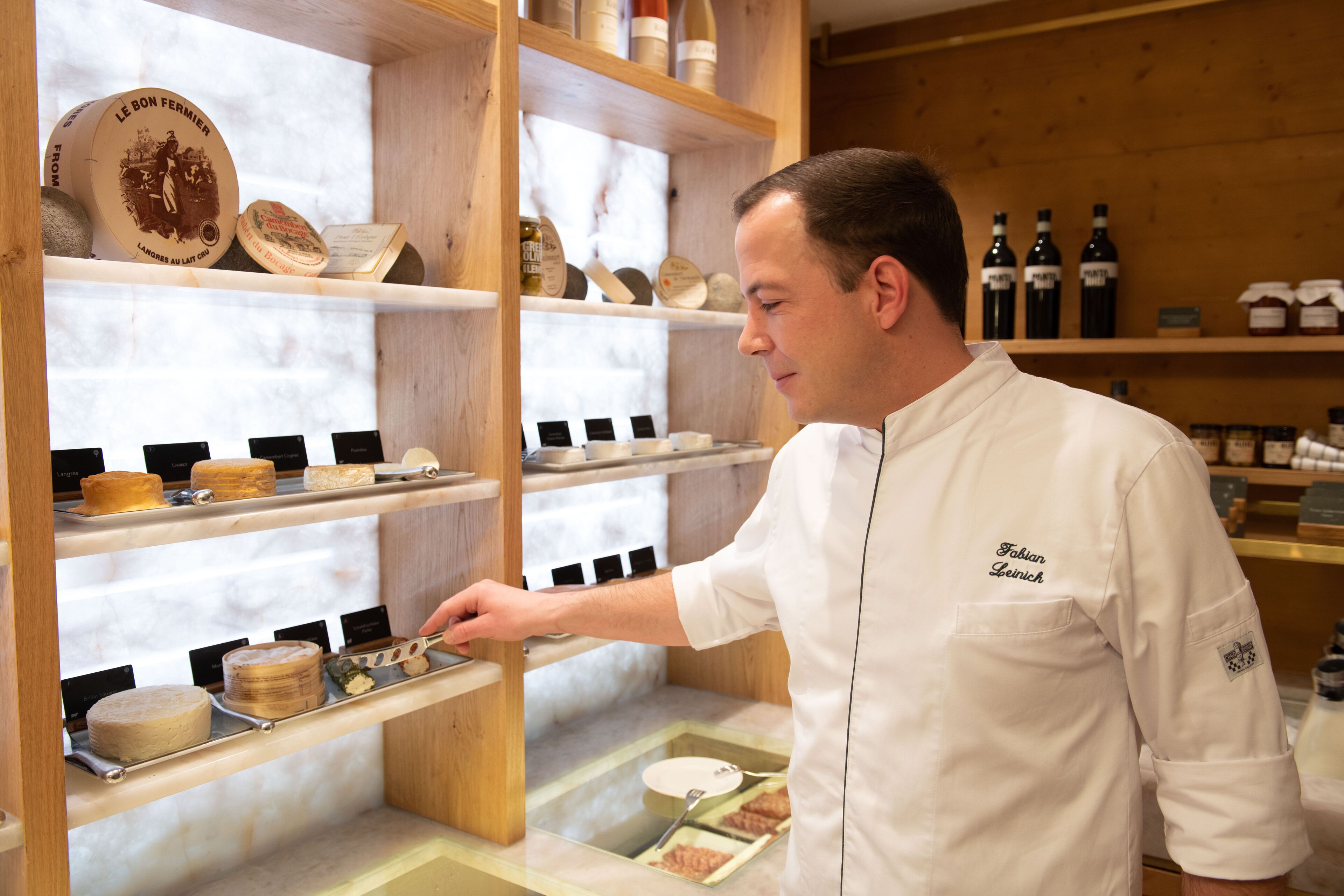 Un chef vêtu d’un uniforme blanc, avec l’inscription « Fleisch » sur la poitrine, tend la main vers un plateau rempli de différents plats, qui se trouve sur une étagère en bois.