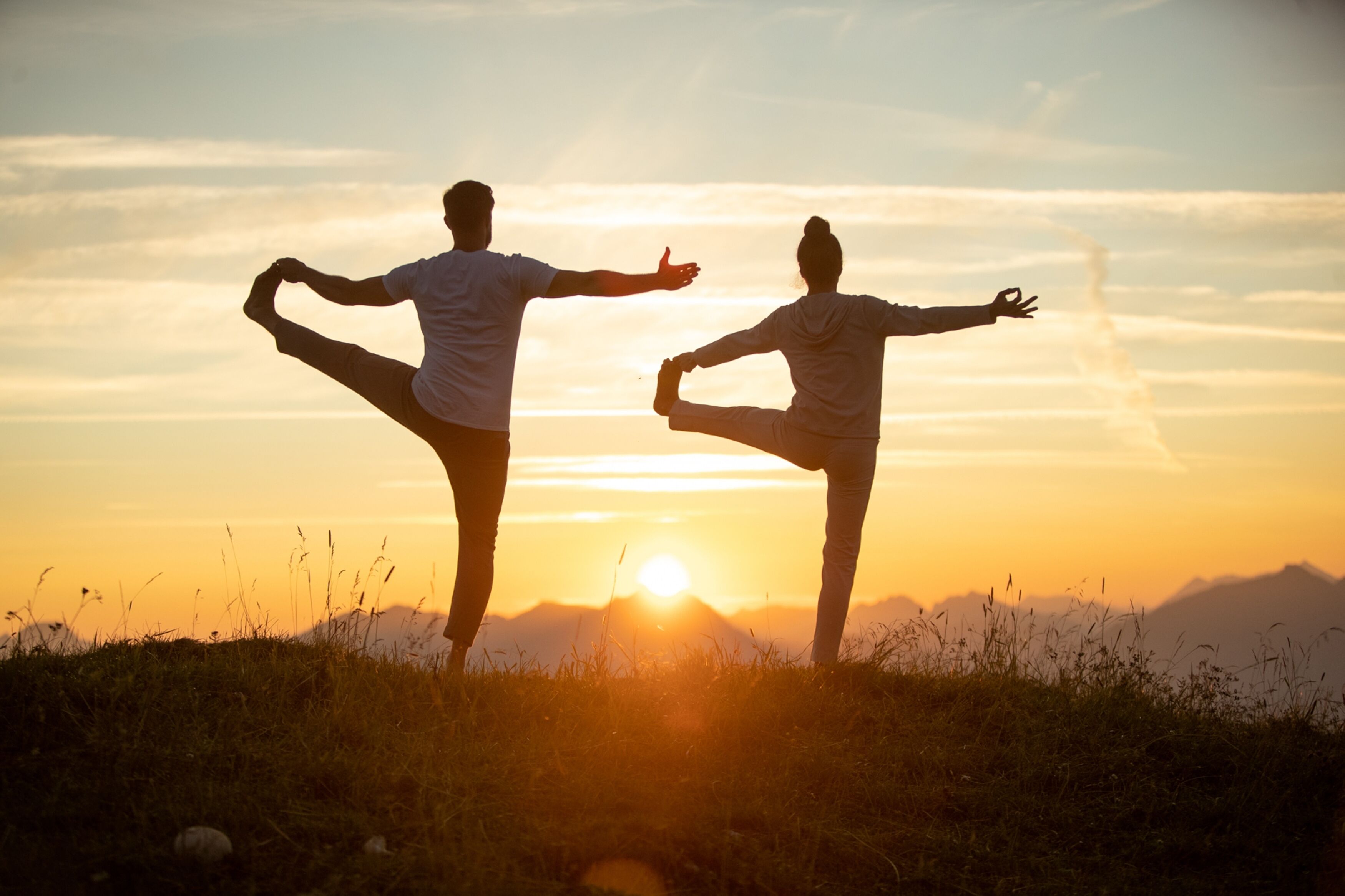 Dos personas realizando posturas de yoga en un campo cubierto de hierba con un atardecer de fondo.