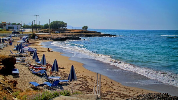A beach with rows of sun loungers and umbrellas, a rocky shoreline, and a clear blue sky. - Beach Front Villa Peri (Hersonissos)