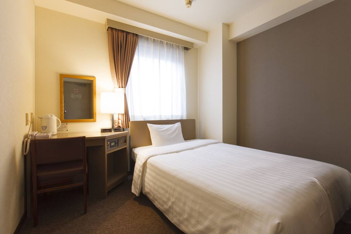 A hotel room with a bed, a desk with a lamp, a telephone, and a window with curtains.