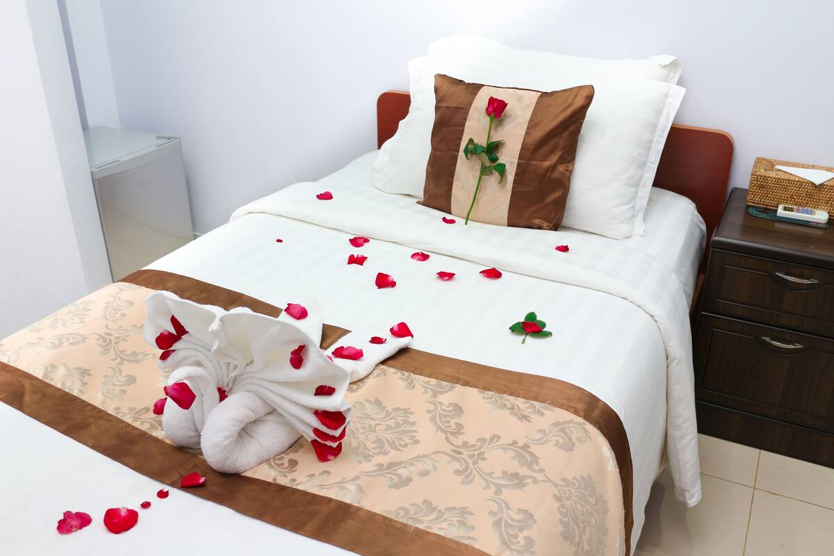 Un lit parfaitement fait, décoré de pétales de rose et surmonté d’une serviette en forme de cygne, un oreiller marron orné d’une rose, et une table de chevet avec un panier et une télécommande.
