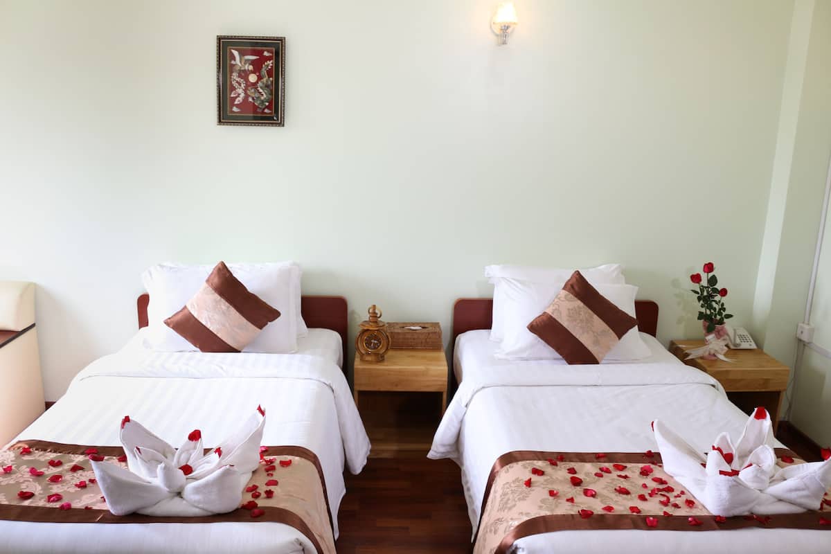 Une chambre d’hôtel avec deux lits, chacun recouvert d’une courtepointe blanche, agrémenté d’oreillers marron et d’une composition florale en serviettes. Une table de chevet en bois se trouve entre les deux lits, sur laquelle reposent une horloge et une petite plante.