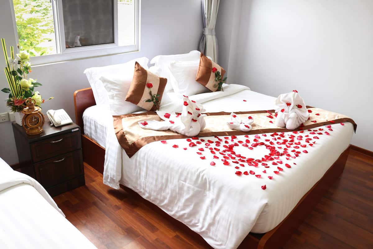 Un lit bien fait, décoré de pétales de roses et agrémenté d’une sculpture en serviette, une table de chevet avec un téléphone et un vase de fleurs, et une fenêtre avec des rideaux.