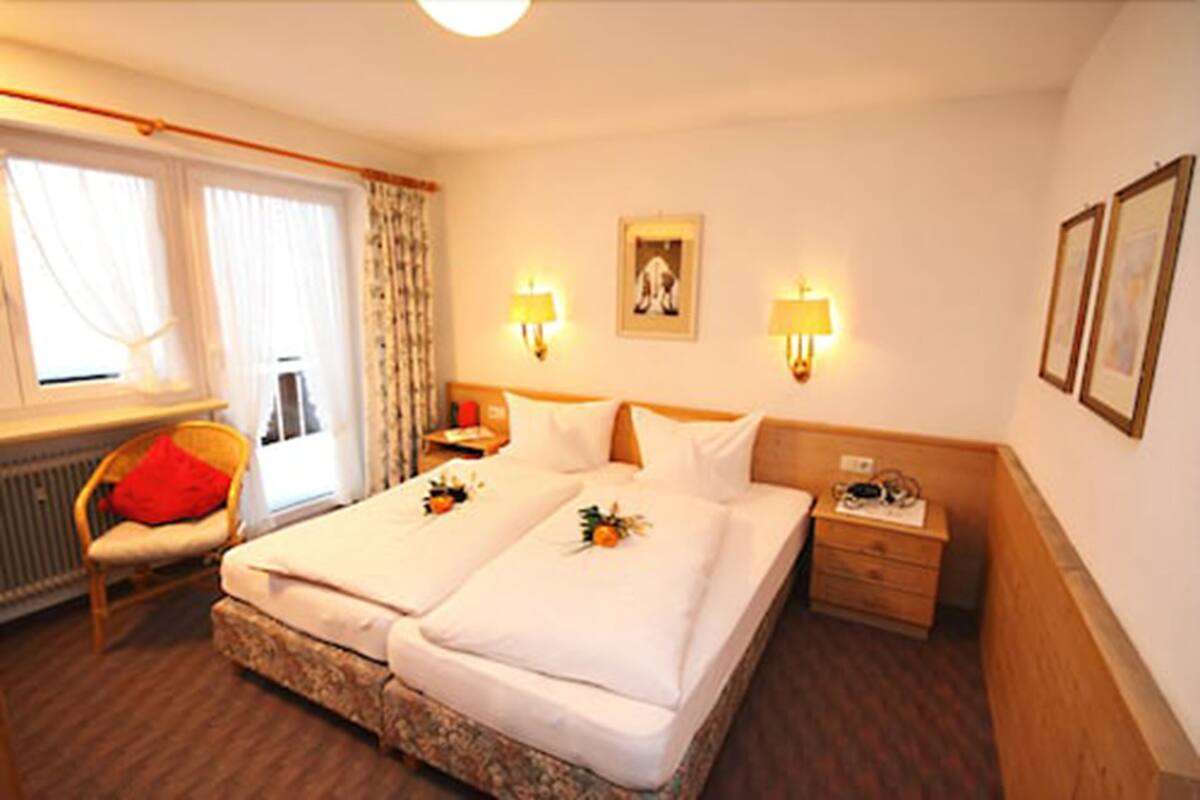 A double bed with white linens, two bedside tables, a nightstand, a chair, and a window with curtains.