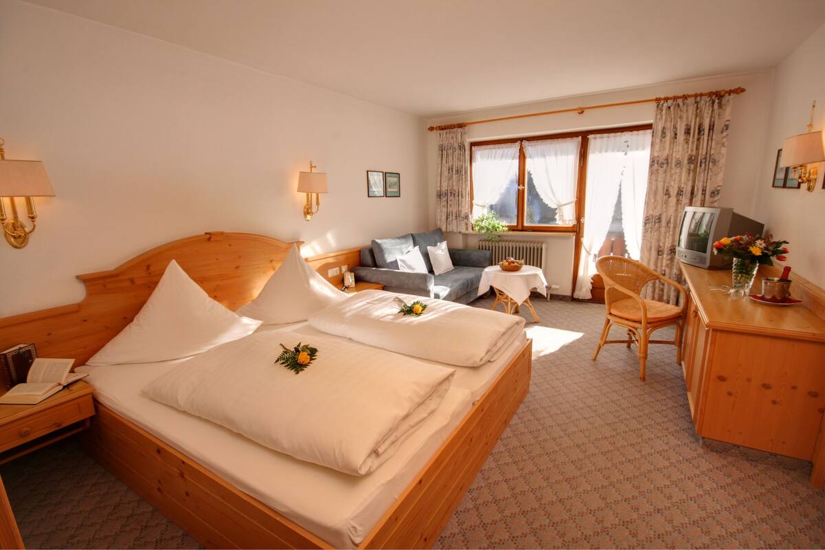 A hotel room with a wooden bed, a sofa, a desk with a TV, and a view of a mountain through the window.