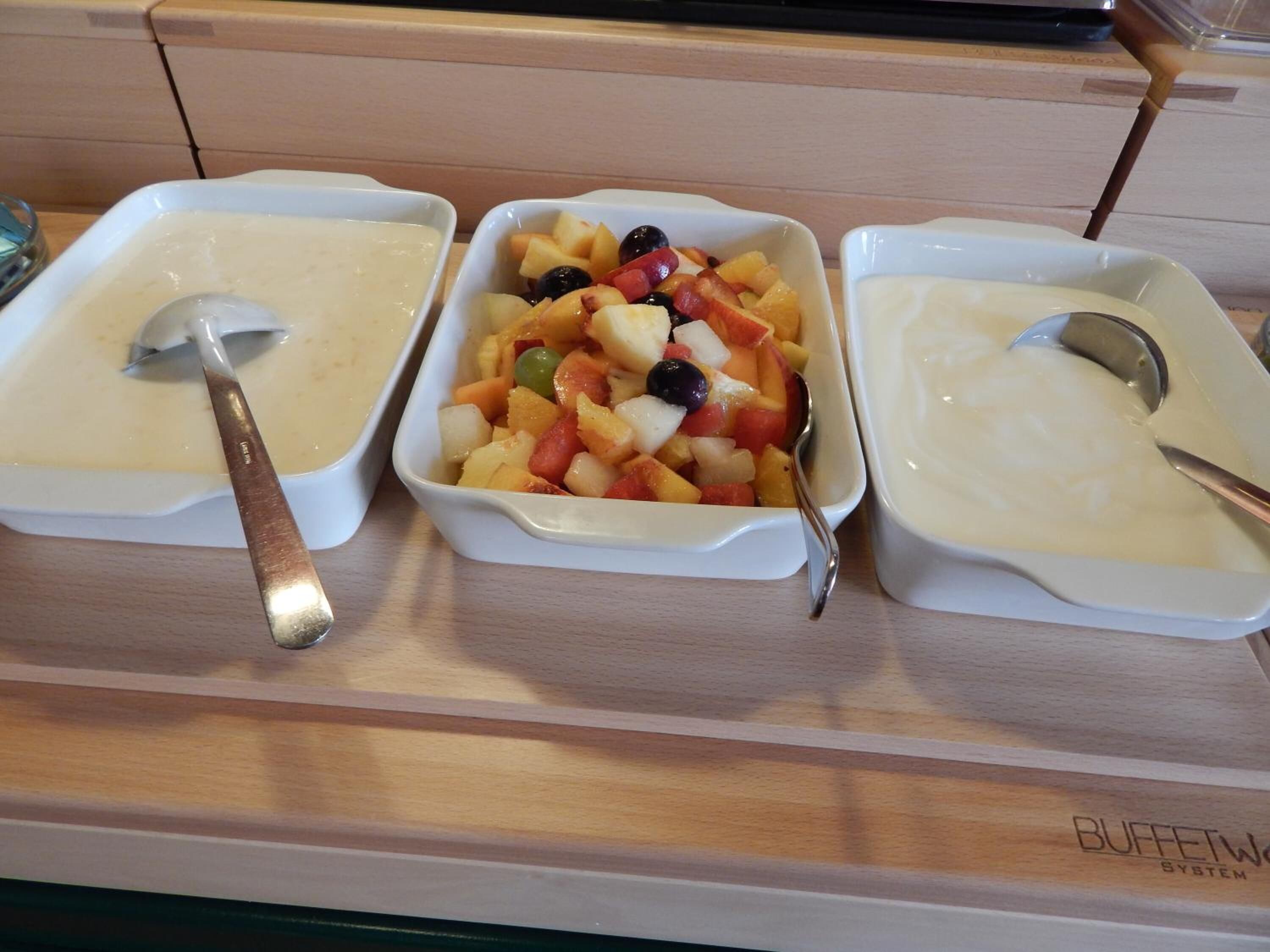 Drei weiße Schüsseln, jeweils mit einem Löffel, gefüllt mit einer cremigen Masse und Obstsalat.