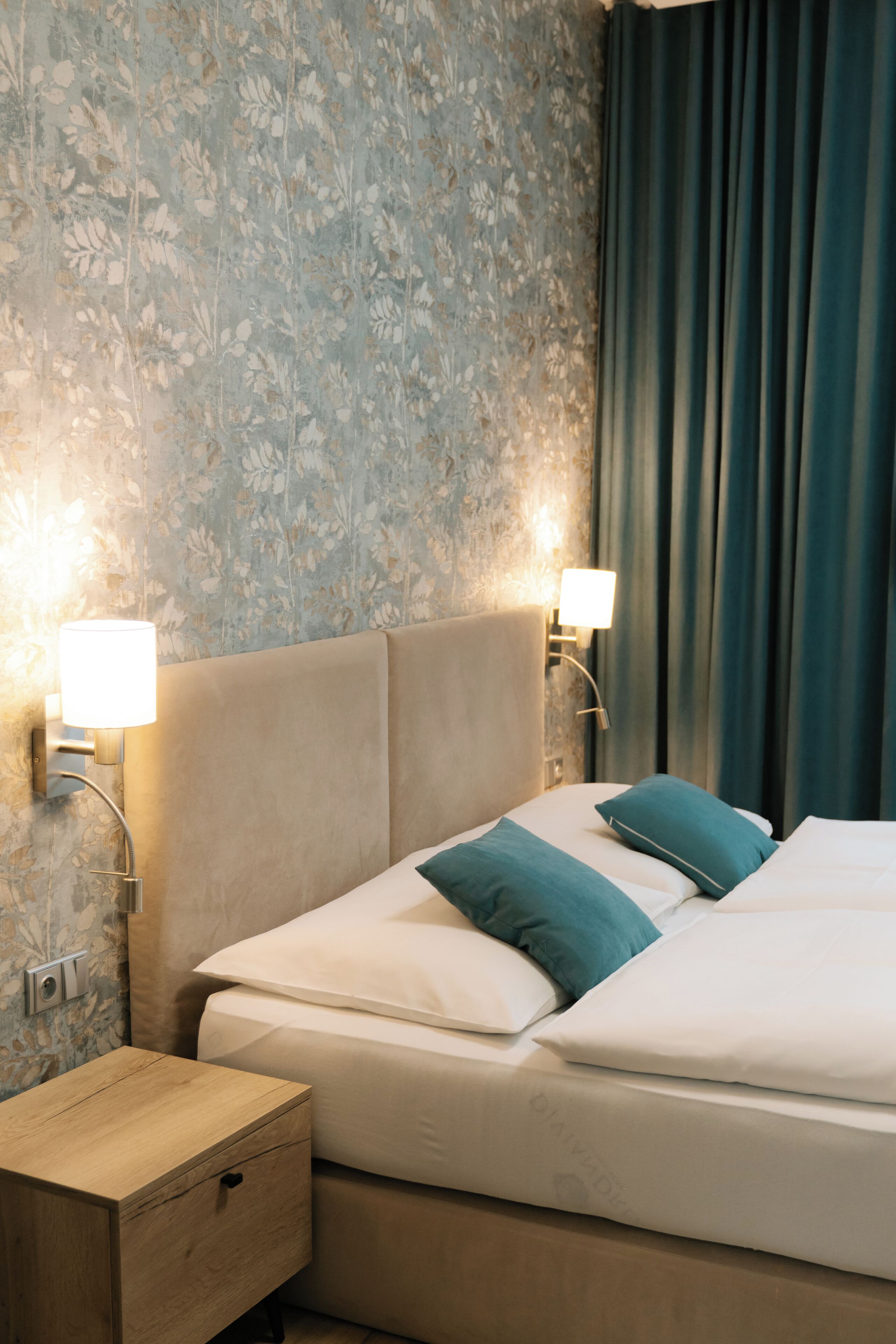 A neatly made bed with white and teal bedding, a wooden nightstand with a drawer, and a wall with floral wallpaper.