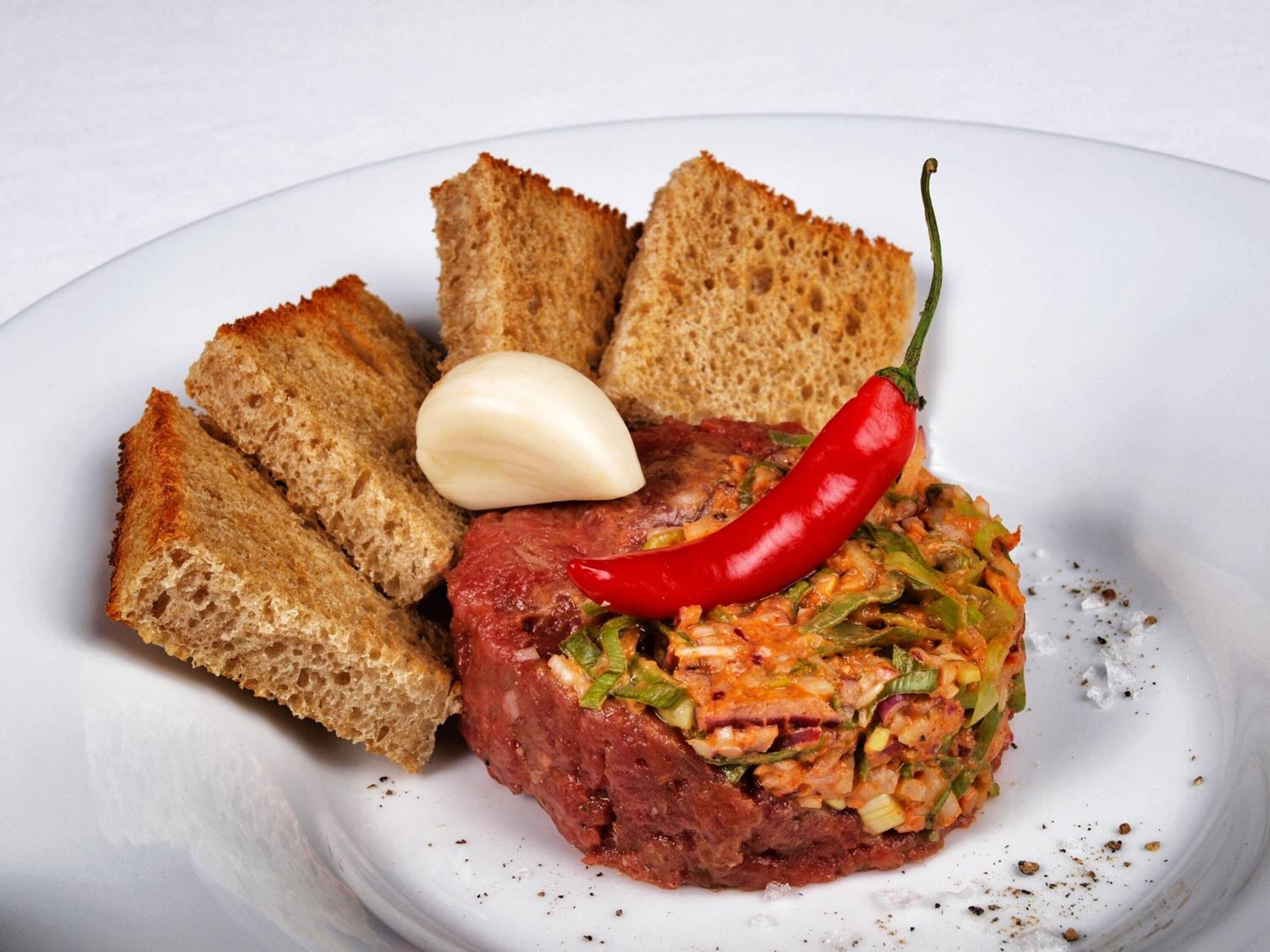 Ein Teller mit einem Fleischgericht, einer roten Chilischote, einer Knoblauchzehe und geröstetem Brot.