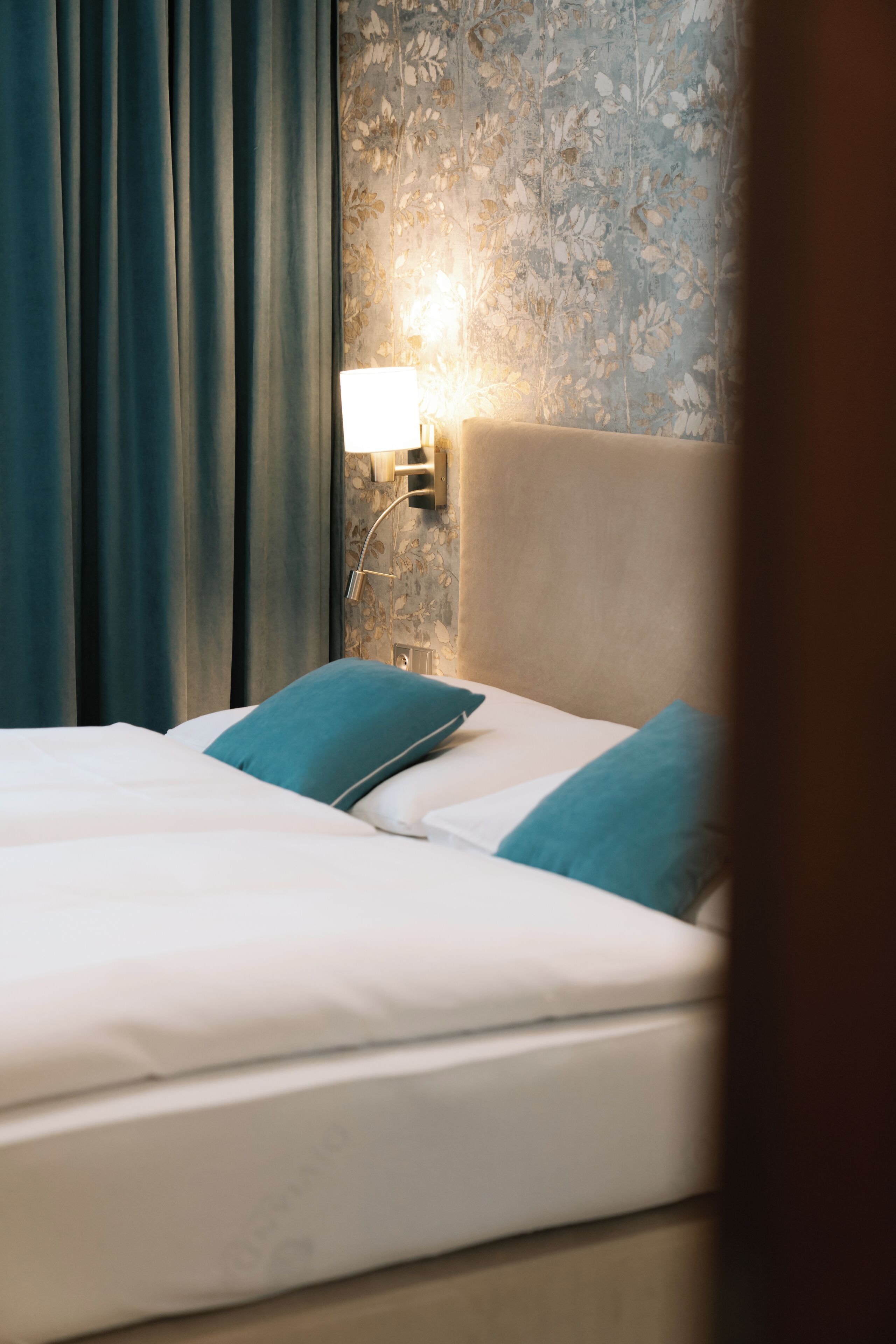 Una cama bien hecha con sábanas blancas y almohadillas color turquesa, una lámpara de pared y un estampado de papel tapiz floral.