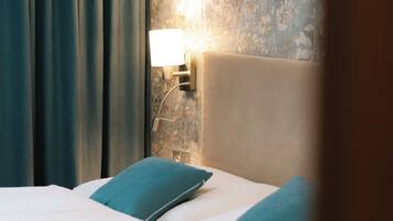 Una cama bien hecha con sábanas blancas y almohadillas color turquesa, una lámpara de pared y un estampado de papel tapiz floral.