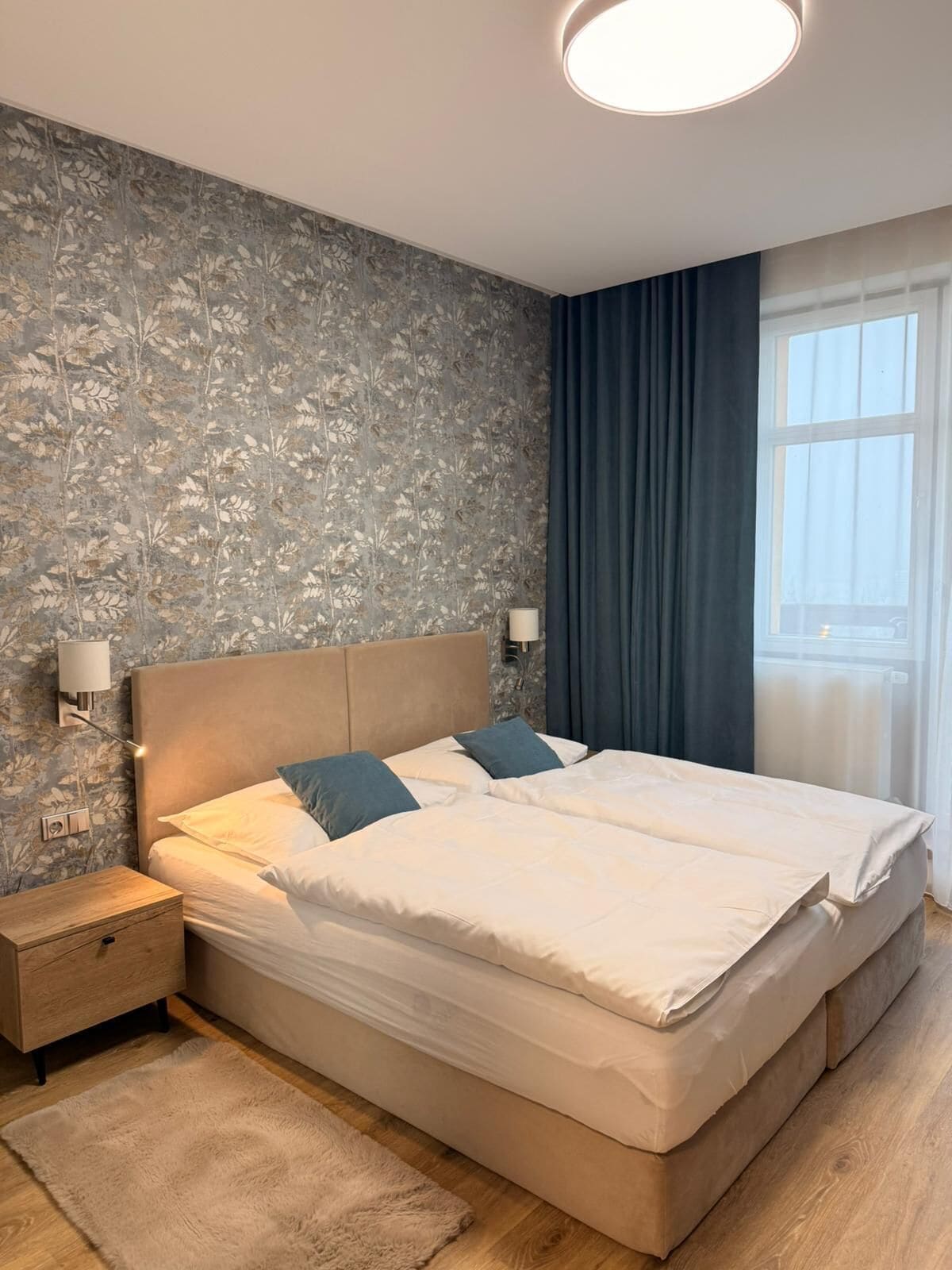 A double bed with a headboard, two bedside tables, a fluffy rug, and a window with curtains.