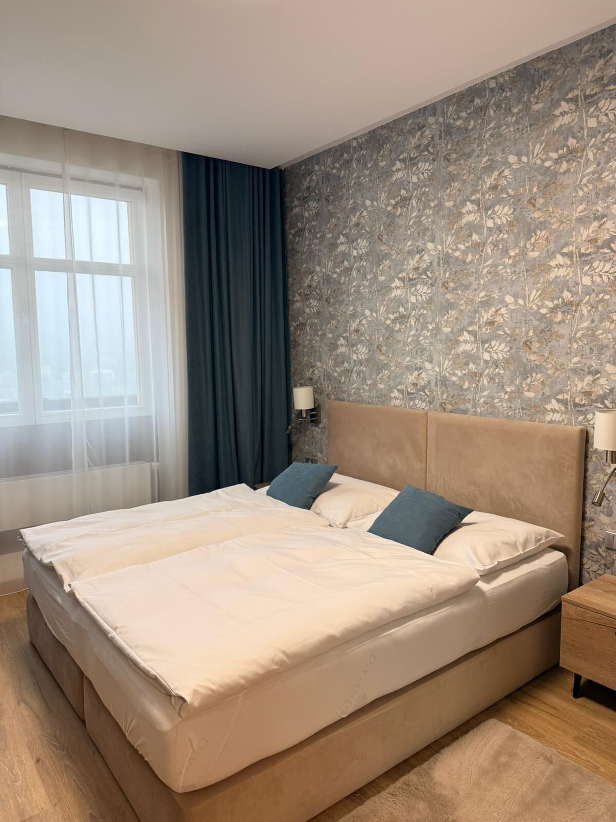 Una cama grande con cabecera color beige, dos almohadas y una mesita de noche de madera con una lámpara.