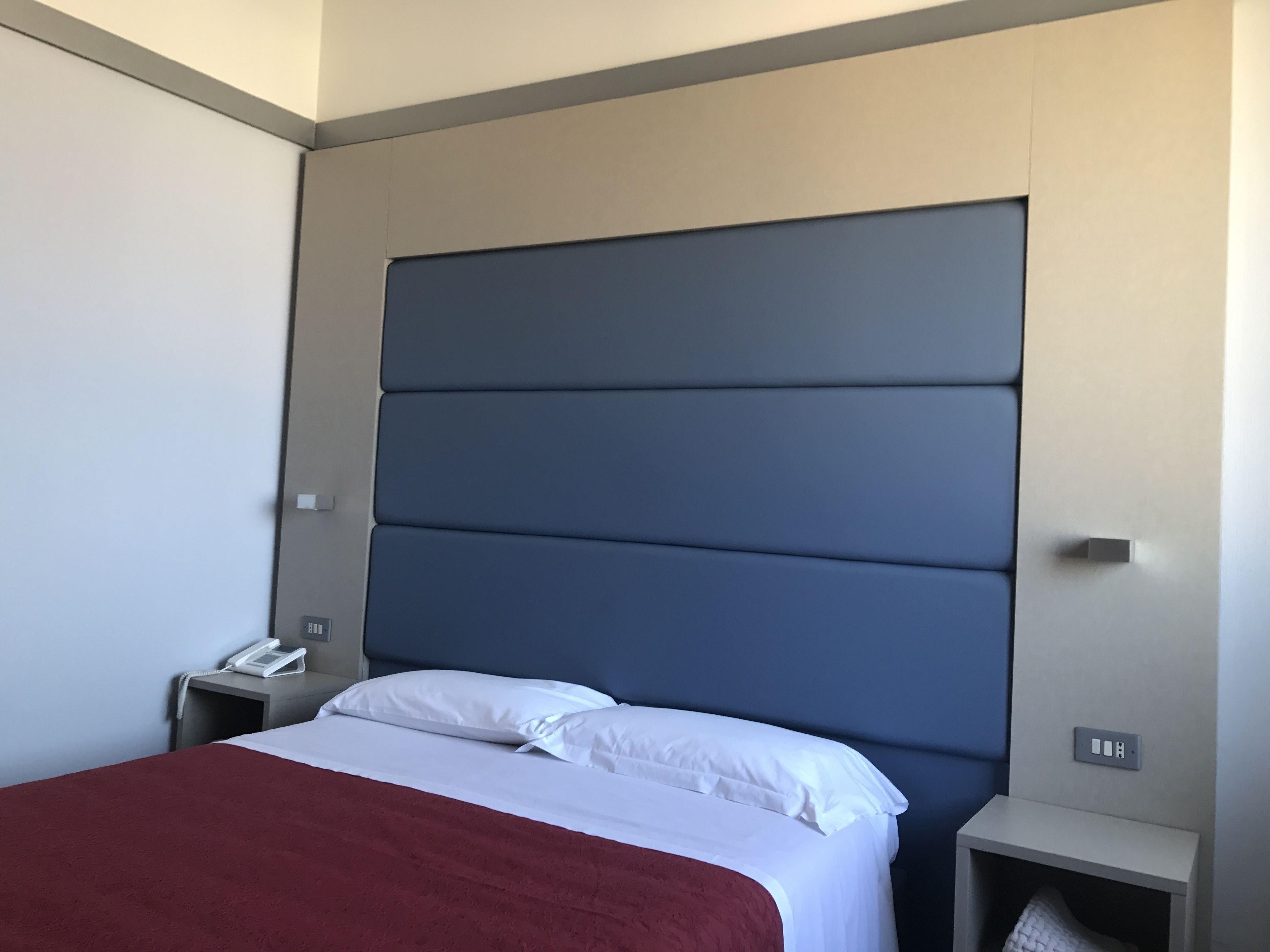 Una camera d'albergo moderna con un letto grande, lenzuola bianche, una coperta rossa e un comodino con il telefono.