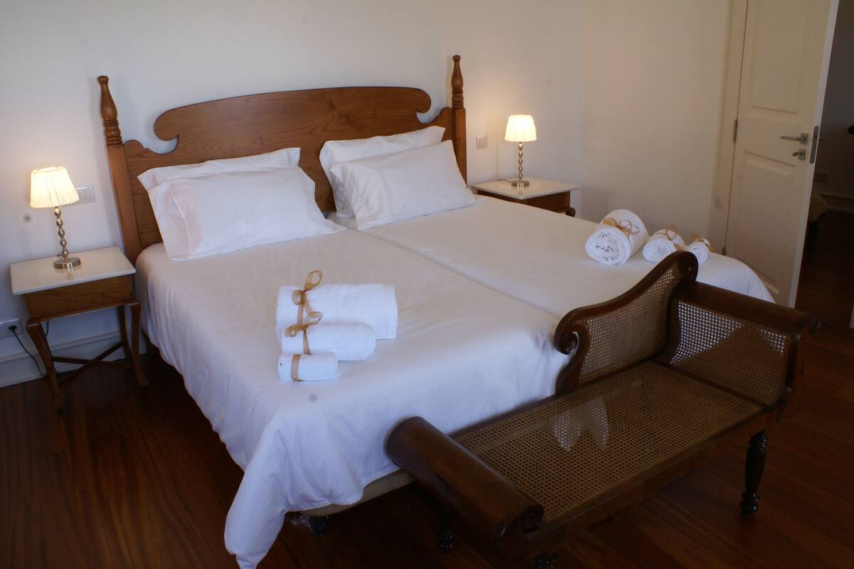 A neatly made bed with white linens and two pillows, a wooden bench, and two bedside lamps.