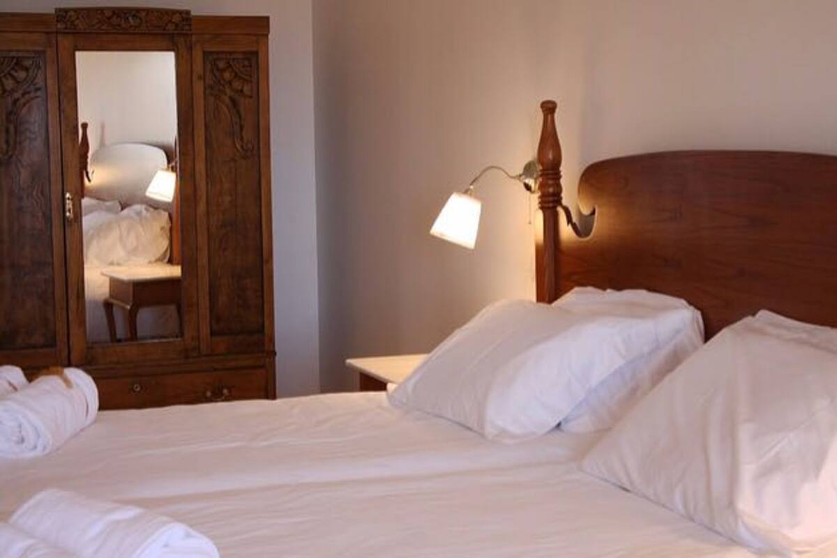 A neatly made bed with white linens and pillows, a wooden headboard, a bedside lamp, and a wooden wardrobe with a mirror.