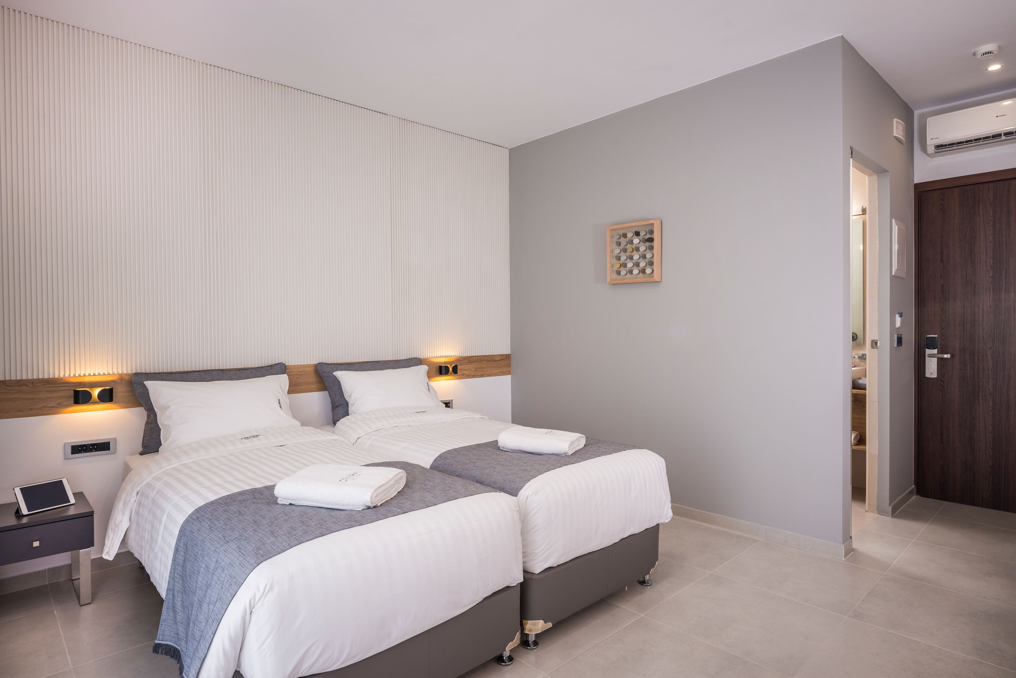 A modern hotel room with two beds, a bedside table with a tablet, a wall-mounted light, and a wooden door.