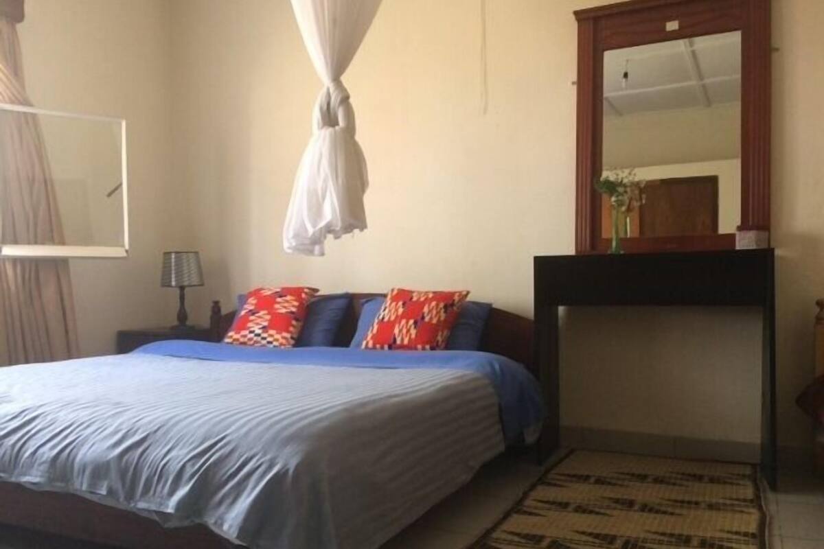 A bedroom with a bed, a nightstand, a mirror, and a window with curtains.
