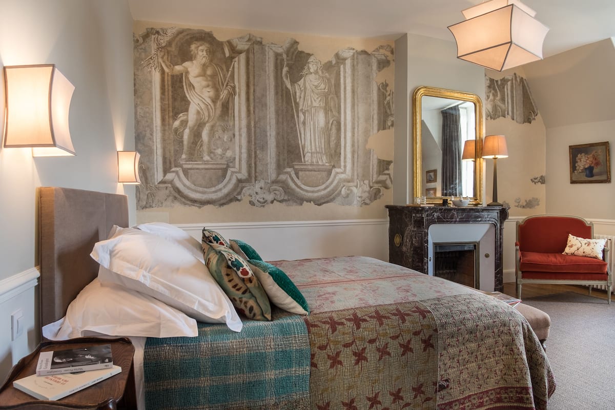 Une chambre d’hôtel avec un grand lit, un bureau avec une lampe, une chaise, une cheminée et un mur orné d’une fresque.