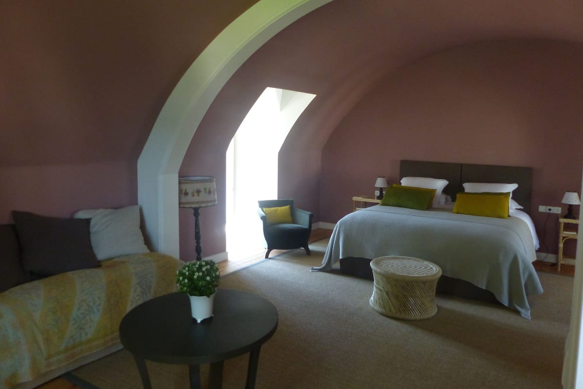 Une chambre confortable avec un plafond cintré, un lit agrémenté de plusieurs coussins, une chaise, une petite table avec une plante et une lampe.