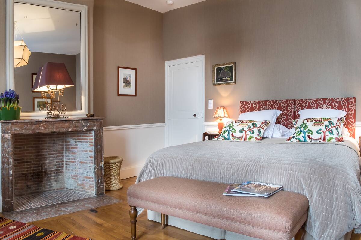 Une chambre avec une cheminée en briques, un grand lit agrémenté d’oreillers à motifs et un banc avec un livre.