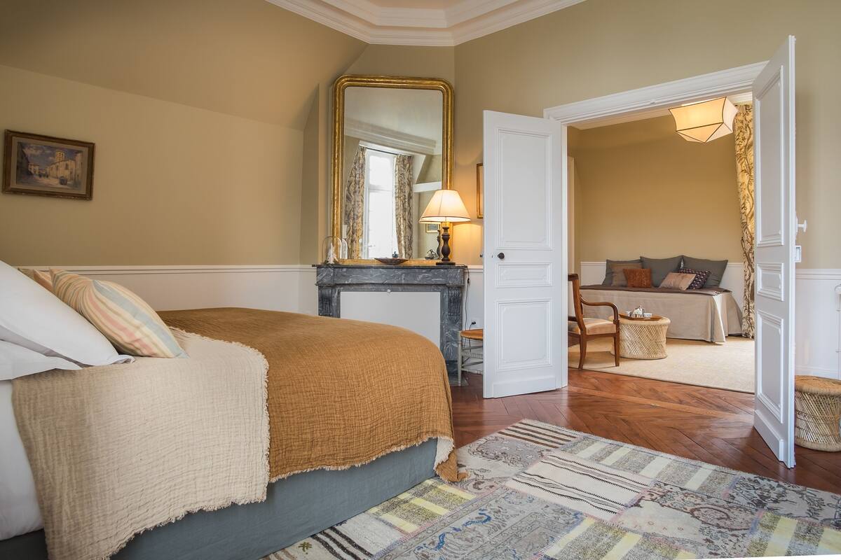 Une chambre à coucher comprenant un lit, une cheminée, un miroir et une porte donnant sur un coin salon avec un canapé et une lampe.