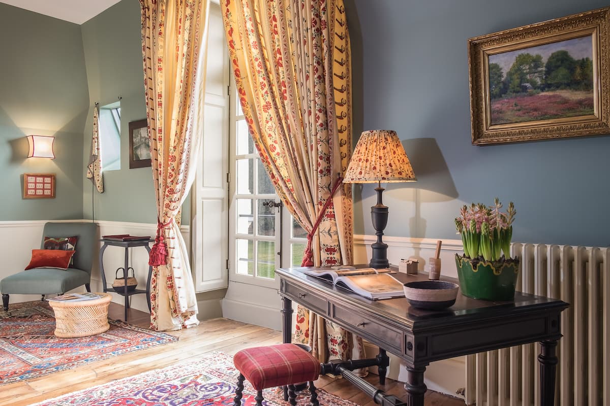 Une pièce chaleureuse avec un plancher en bois, un fauteuil à carreaux rouges, une table avec une lampe, un tableau encadré et une fenêtre avec des rideaux.