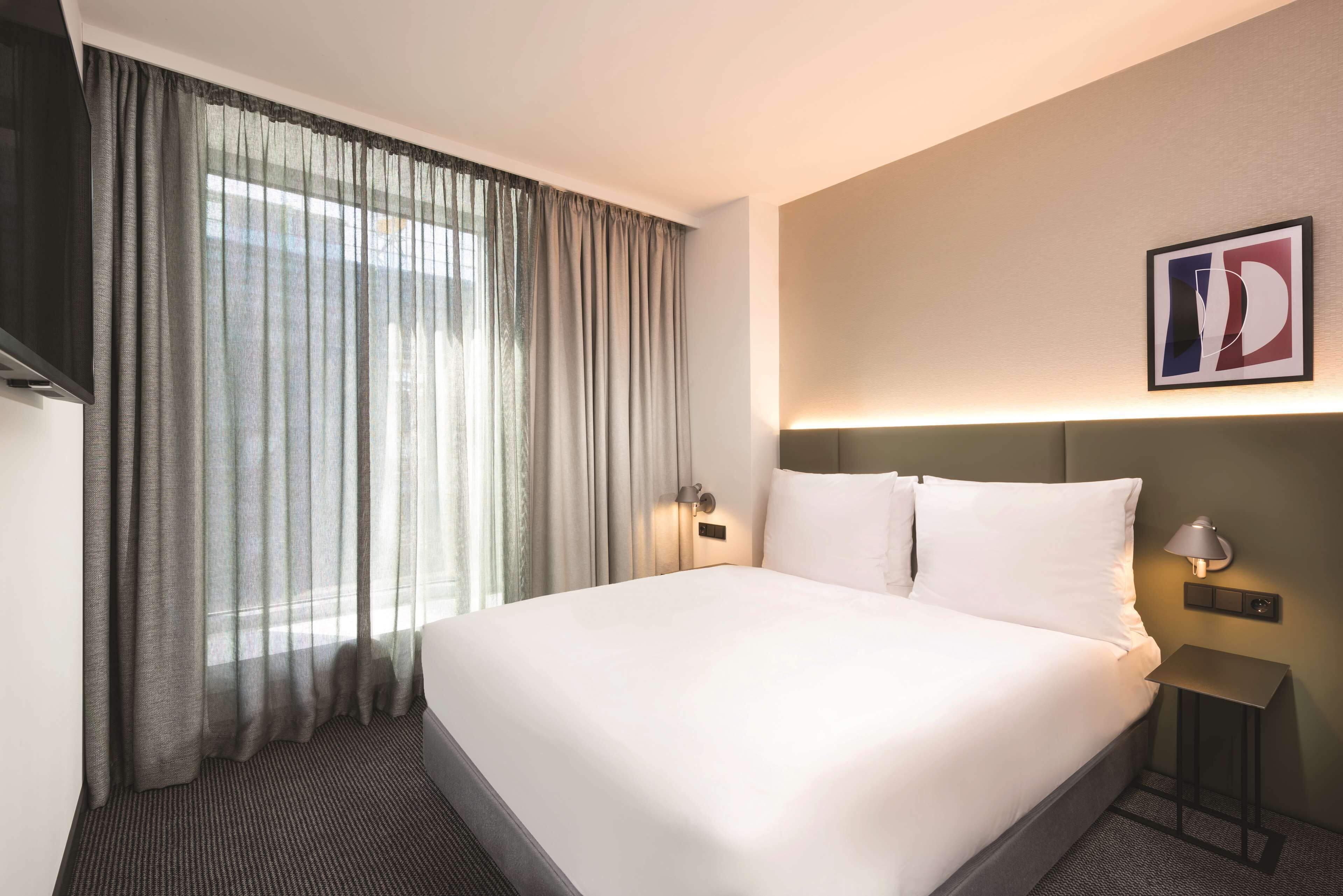 A hotel room with a large bed, a flat-screen TV, a bedside table with a lamp, and a window with sheer curtains.