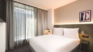 A hotel room with a large bed, a flat-screen TV, a bedside table with a lamp, and a window with sheer curtains.