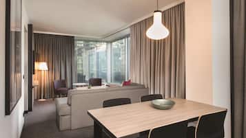 A modern hotel room with a sofa, dining table, and chairs. There is a window with curtains, a lamp, and a view of buildings outside.