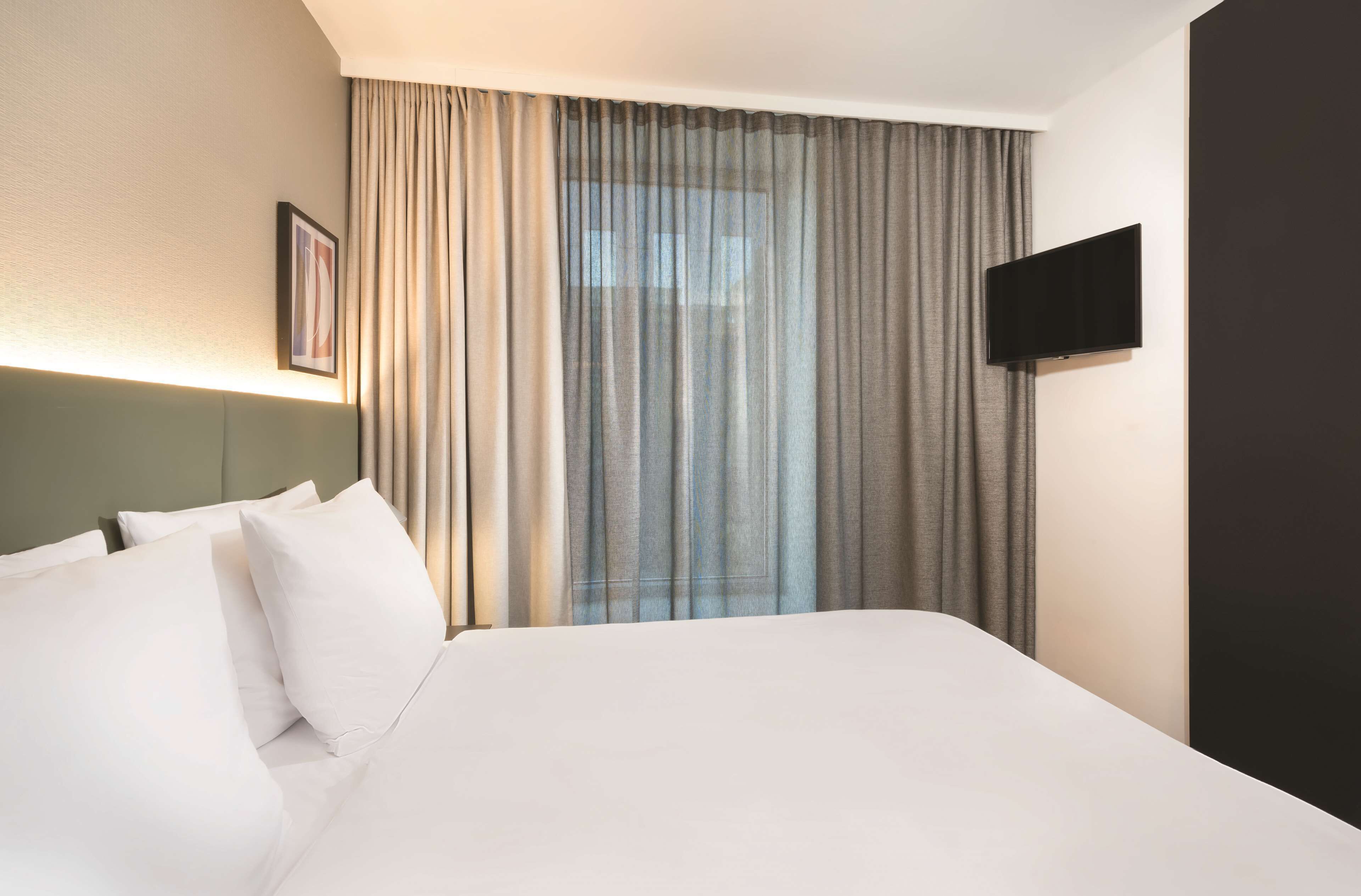 Ein Hotelzimmer mit einem großen Bett, einem flachbildfernseher an der Wand und einem Fenster mit transparenten Vorhängen.