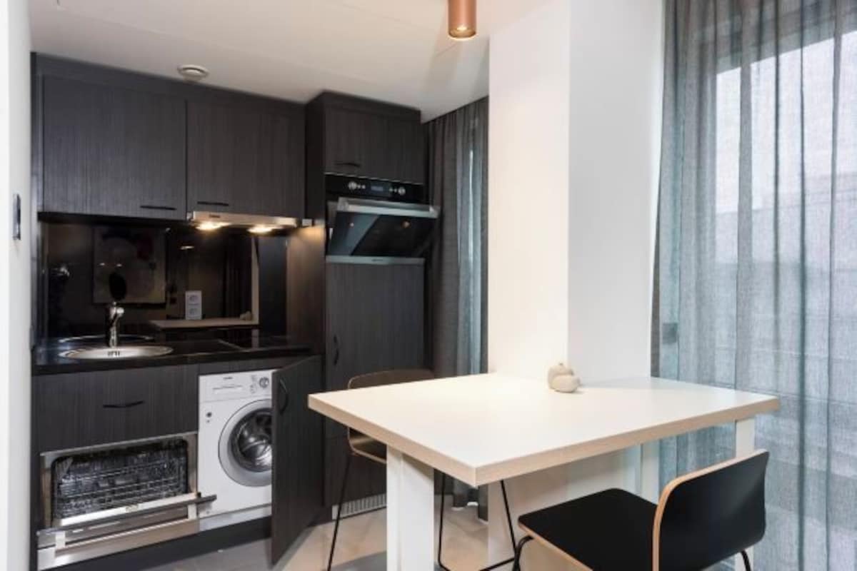 A compact kitchen with dark cabinetry, a built-in oven, a dishwasher, and a small dining area with a white table and two chairs.