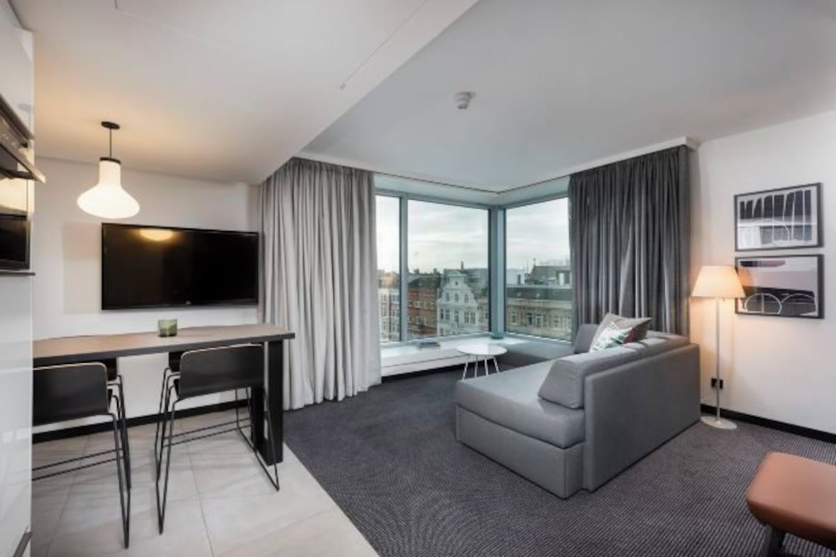A modern hotel room with a large window, a grey sofa, a dining area with a TV, and a lamp.